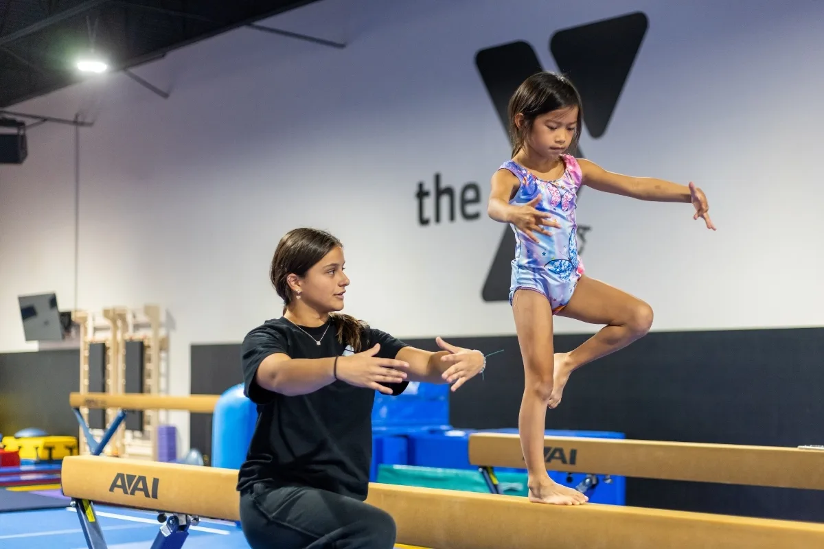 Gymnastics coach helping a child on a balance beam inside a gymnastics facility