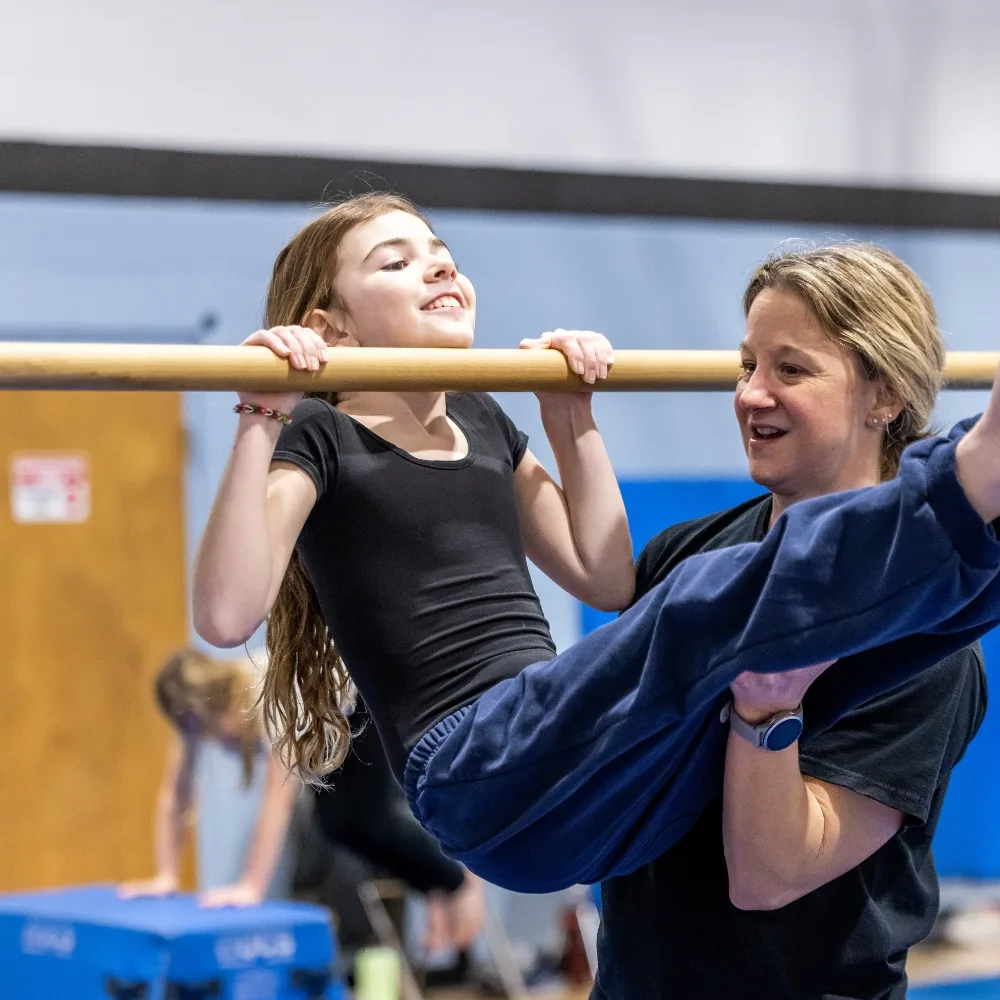 Gymnastics trainer helping youth gymnastics athlete on uneven bar at indoor gymnastics facility